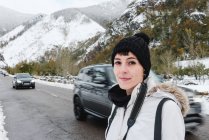 Femme avec piercing nez en chapeau noir et veste d'hiver blanche debout sur route asphaltée avec des montagnes enneigées sur fond — Photo de stock