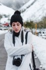 Mulher de casaco de inverno branco com capuz e calças pretas andando no meio da estrada de asfalto entre montanhas nevadas com câmera — Fotografia de Stock