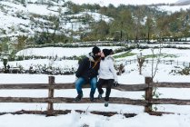 Романтичний чоловік і жінка в зимовому одязі цілуються, сидячи на маленькому дерев'яному паркані на природі під час сніжної погоди — стокове фото