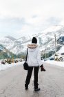 Visão traseira de pessoa em jaqueta de inverno branca com capuz na cabeça em pé no meio da estrada de asfalto a pé de montanhas em tempo nevado — Fotografia de Stock