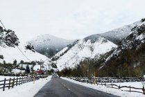 Route asphaltée entre montagnes enneigées et forêts avec petites clôtures en bois de village sur les côtés pendant la journée d'hiver — Photo de stock