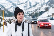 Femme marchant au milieu de la route asphaltée entre les montagnes enneigées avec des voitures sur le fond — Photo de stock