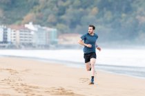 Atleta masculino barbudo em desgaste ativo correndo durante praia vazia de areia com montanhas verdes em fundo embaçado — Fotografia de Stock