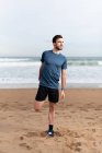 Hombre deportivo en desgaste activo estirando las piernas para correr en la playa de arena vacía y mirando hacia otro lado - foto de stock