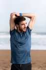 Bearded male athlete in blue t shirt stretching arms and looking away with sandy seashore on blurred background — Stock Photo