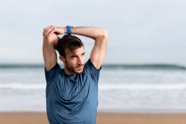 Bearded male athlete in blue t shirt stretching arms and looking away with sandy seashore on blurred background — Stock Photo