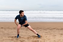 Gymnaste masculin en vêtements de sport étirant les jambes et regardant loin sur la plage de sable vide avec mer bleue et ciel sur fond flou — Photo de stock