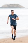 Бородатий спортсмен в активному носінні бігає під час порожнього піщаного пляжу з зеленими горами на розмитому фоні — стокове фото