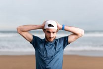 Sportsman in t shirt blu con le mani dietro la testa sul cappuccio bianco guardando la fotocamera con spiaggia sabbiosa vuota su sfondo sfocato — Foto stock