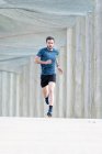 Bearded male athlete in blue t shirt and shorts running outdoors under cover looking at camera — Stock Photo