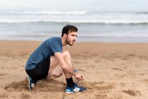 Vista lateral del hombre deportivo barbudo en desgaste activo sentado en cuclillas y atando cordones en la playa vacía de arena y mirando hacia otro lado - foto de stock