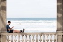 Вид збоку людини в активному одязі, що сидить на кам'яному паркані і спирається на колону під час використання мобільного телефону — стокове фото