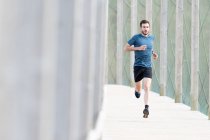 Hochmotivierte bärtige männliche Athleten in blauem T-Shirt und kurzen Hosen laufen im Freien unter Deckung in die Kamera — Stockfoto