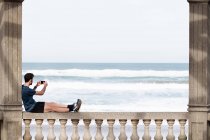 Vista lateral del hombre sin rostro en desgaste activo sentado en la valla mecedora y apoyado en la columna mientras toma una foto del mar azul en el teléfono móvil - foto de stock