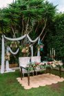 Ensemble pour cérémonie de mariage avec table de jeunes mariés décorée avec arrangement de fleurs et placée sur le tapis contre l'arbre tropical vert avec arc de mariage blanc — Photo de stock