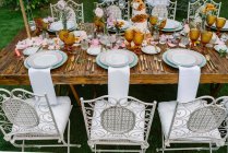 Mesa de casamento de madeira sem pano decorado com pétalas de flores e buquês simples em vasos e servido com pratos e copos de laranja e cadeiras brancas rústicas ao redor — Fotografia de Stock