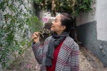 Jeune femelle vêtue de vêtements décontractés touchant des plantes vertes, sentant des fleurs et souriant dans un couloir rocailleux — Photo de stock