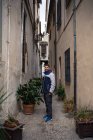Молодий чоловік у відпустці в повсякденному теплому одязі, що стоїть на вузькій старій вулиці з горщиками із зелених рослин — стокове фото