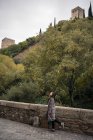 Вид збоку на жінку-мандрівника в казуальній шубці, що стоїть на старій мосту з бруківкою з парканом і зеленими пагорбами з стародавнім замком — стокове фото