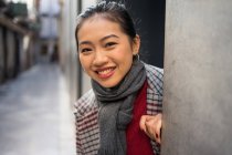 Overjoyed Asian female in casual coat peering from wall of modern building and looking in camera at Albaicin in Granada, Spain — Stock Photo