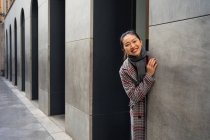 Радостная азиатская женщина в случайном пальто, выглядывающая со стены современного здания и смотрящая в камеру на Альбайсин в Гранаде, Испания — стоковое фото