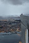 Обратный вид безликих путешественников, стоящих на крыше современного здания и созерцающих удивительный вид вечернего города, расположенного вдоль береговой линии в долине против снежного высокогорья — стоковое фото