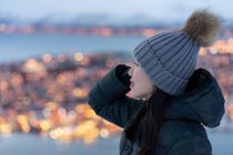 Emocionada joven hembra en chaqueta de plumón caqui y sombrero gris cálido mirando hacia otro lado y contemplando la increíble vista de invierno de la ciudad ubicada en la costa por la noche - foto de stock