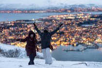 Happy adult man in warm clothes throwing snow like child and enjoying winter while resting with wife together on outskirts standing on hill against amazing view of blurred golden lights of city located on island — Stock Photo