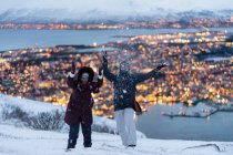 Happy adult man in warm clothes throwing snow like child and enjoying winter while resting with wife together on outskirts standing on hill against amazing view of blurred golden lights of city located on island — Stock Photo