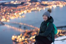 Jovem mulher entusiasmada em casaco cáqui e chapéu cinza quente olhando para a câmera e contemplando incrível vista de inverno da cidade localizada na costa à noite — Fotografia de Stock