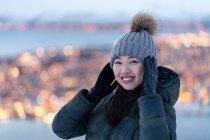 Aufgeregte junge Frau in khakifarbener Daunenjacke und grauem, warmem Hut, die in die Kamera schaut und am Abend erstaunliche Winterbilder von der Stadt an der Küste betrachtet — Stockfoto