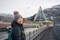Attractive adult Asian woman in warm clothes with backpack smiling at camera while standing on street against blurred exterior of amazing triangle shaped church and snowy hills in Norway — Stock Photo