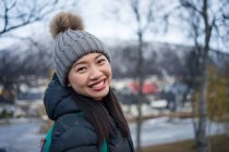 Attrayant adulte asiatique femme en vêtements chauds avec sac à dos souriant à la caméra tout en se tenant dans la rue contre l'extérieur flou de l'étonnante église en forme de triangle et collines enneigées en Norvège — Photo de stock