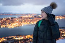 Jeune femme excitée en duvet kaki et chapeau chaud gris regardant loin et contemplant une vue hivernale incroyable de la ville située sur la côte en soirée — Photo de stock