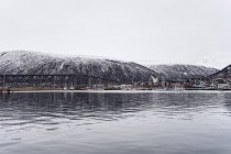 Desde arriba de la bahía tranquila con la ciudad en la costa ordenada poderosa montaña nevada con cielo nublado en el fondo en Tromso en Noruega - foto de stock