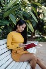 Side view of brunette Asian woman reading book in red cover sitting on white bench in park — Stock Photo
