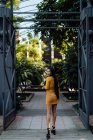 Visão traseira da moda mulher asiática na moda andando ao longo da cerca decorativa de metal e olhando para a câmera sobre o ombro — Fotografia de Stock