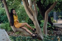 Азійська жінка з довгим волоссям зручно розмістила на стовбурі дерева розтягуючу ногу і розмовляла по мобільному телефону в парку. — стокове фото
