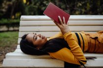 From above of brunette Asian woman reading book in red cover lying down on white bench in park — Stock Photo