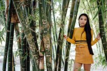 Femme asiatique avec de longs cheveux foncés en chemise jaune et jupe courte debout dans un beau jardin et regardant la caméra — Photo de stock