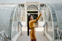 Vista lateral de la mujer asiática en falda corta sonriendo y tomando selfie en teléfono inteligente en vidriosa sala soleada arquitectónica del edificio - foto de stock