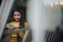 Elegante graciosa sorridente mulher asiática surfar telefone celular e olhando para a câmera na estrada perto de construção de metal — Fotografia de Stock