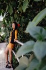 Vista laterale della donna asiatica alla moda con lunghi capelli scuri appoggiati sulla recinzione in metallo con piante rampicanti e guardando la fotocamera — Foto stock