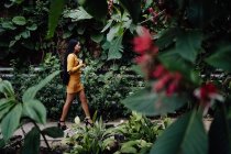 Vue latérale de la femme asiatique à la mode avec de longs cheveux foncés en chemise jaune et jupe courte marchant dans un beau jardin et regardant loin — Photo de stock