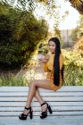Affascinante donna asiatica alla moda con lunghi capelli scuri in camicia gialla guardando la fotocamera e mettendo lucidalabbra nel parco soleggiato — Foto stock