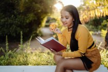 Bruna asiatico donna lettura libro in rosso copertina e mangiare gustoso mela seduta su bianco panchina in parco — Foto stock