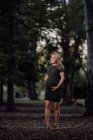 Happy pregnant woman in casual dress stroking belly while standing on pathway in park with green trees — Stock Photo