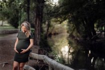 Schöne schwangere Frau in lässigem Kleid, lächelnd und Händchen haltend auf dem Bauch, während sie neben Holzbrücke steht — Stockfoto
