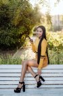 Side view of stylish brunette Asian woman with smartphone touching high heel shoes sitting on back of white bench — Stock Photo