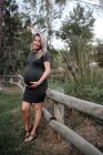 Mulher grávida bonita em vestido casual sorrindo e de mãos dadas na barriga enquanto está ao lado da ponte de madeira — Fotografia de Stock
